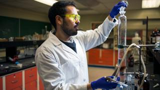 A male student in a chemistry lab is pouring fluid from one beaker into a larger test tube.